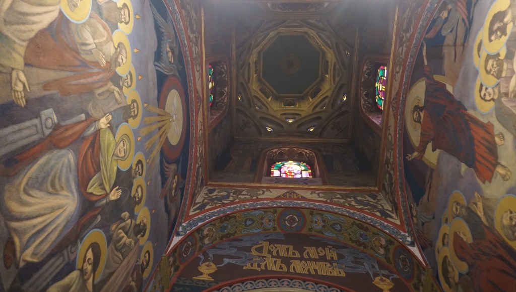 Interior view of a church showcasing colorful frescoes on the walls and a decorative ceiling with stained glass windows.